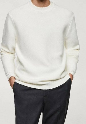 Homme portant un pull en maille blanc uni avec des poignets et un ourlet côtelés, associé à un pantalon noir, les mains dans les poches.