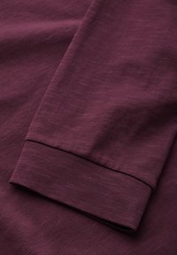 Maroon pitkähihaisen paidan kangas, jossa on sileä rakenne. Paidassa on kääritty hiha yksinkertaisella helmitäytteellä ja hieman heatheroitunut ulkonäkö.