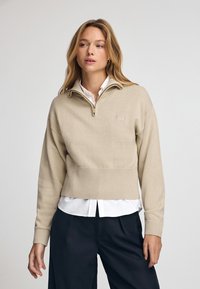 Beżowy cropped sweter z półzapinanym kołnierzem, wykonany z teksturowanej tkaniny i wykończony ściągaczem. Noszony na białej koszuli z kołnierzykiem.