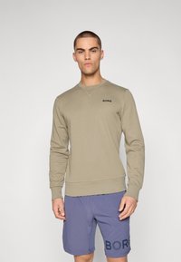 Olive sweatshirt with a crew neck and long sleeves, featuring a subtle black logo. Paired with blue shorts displaying the same brand name.