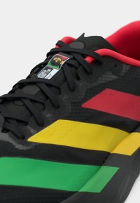 Μαύρο αθλητικό παπούτσι με ρίγες σε κόκκινο, κίτρινο και πράσινο, μαύρα κορδόνια, κόκκινη εσωτερική επένδυση και ένα μικρό γραφικό του Τσε Γκεβάρα στην ετικέτα της γλώσσας.