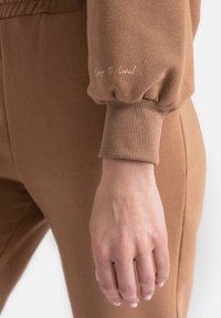 Brązowy dresowy sweter z bawełny z bufkami na rękawach i prążkowanym wykończeniem rękawów, z małym nadrukiem tekstowym, w zestawie z widocznymi brązowymi spodniami.