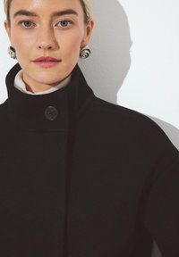 Sort uldfrakke med høj krave, der har en knaplukning. Modellen har en lys turtleneck på under. Sølv spiraløreringe.
