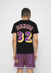 Camiseta de algodón negra con la palabra "JOHNSON" y el número "32" en grandes letras moradas y amarillas en la parte posterior. Combinada con pantalones cortos a rayas moradas.