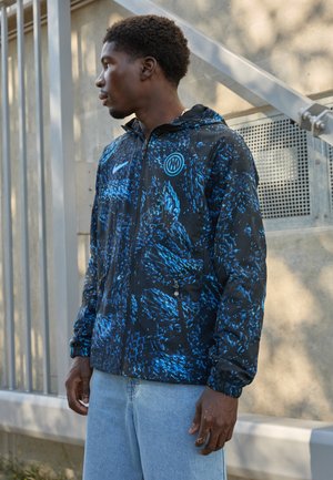 Veste noire avec motif abstrait bleu, fermeture éclair, poignets élastiques et capuche. Arbre avec logo Nike et poches latérales à fermeture éclair. Design décontracté.