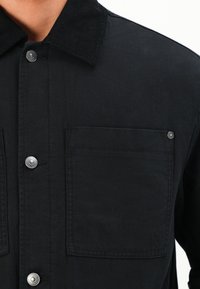 Man wearing a black button-up shirt with a corduroy collar and a square chest pocket with a metal rivet on the top right corner.