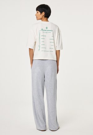 Frau mit kurzen schwarzen Haaren steht mit dem Rücken zur Kamera, trägt ein weißes, kurz geschnittenes T-Shirt mit grünem "Room service"-Schriftzug und hellgraue, weit geschnittene Hose.