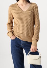 Pull en V beige texturé avec des manches côtelées, associé à un jean bleu foncé et un petit sac à main blanc.