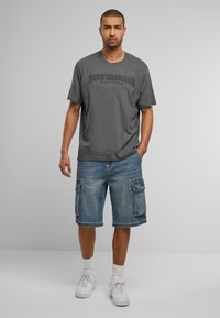 Pilkas medvilninis marškinėliai su "TRUE RELIGION" logotipu, derinami su šviesiai mėlynomis džinsinėmis kargo šortais, turinčiais šonines kišenes ir laisvu siluetu. Balti sportbačiai.