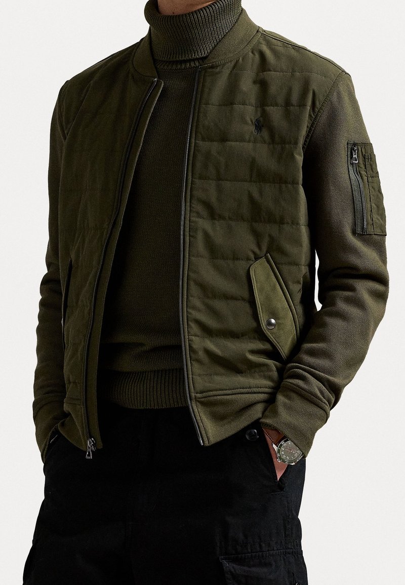 Homme portant une veste bomber matelassée vert olive sur un pull à col roulé assorti, avec un pantalon cargo noir et une montre-bracelet.