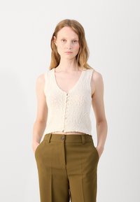 Dzianinowy bezrękawnik w kolorze off-white, z głębokim dekoltem w kształcie litery V, z guzikami z przodu i teksturowanym materiałem. Połączony z oliwkowymi spodniami.