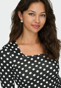Top nero con pois bianchi, scollatura rotonda, texture liscia. Il modello ha capelli mossi e orecchini a bottone in argento, con uno sfondo neutro.