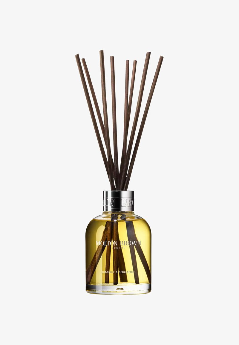 Reed diffuser with clear glass bottle, silver collar, yellow liquid, and eight brown reeds. Label reads "Molton Brown, Orange & Bergamot."