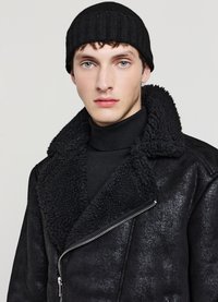 Giacca nera in ecopelle con colletto in shearling, chiusura con zip e finitura strutturata, abbinata a un berretto a coste nero e un dolcevita.