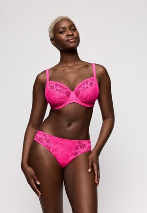 Vrouw met kort blond haar, draagt een felroze kanten beha en bijpassend ondergoed, staand tegen een effen lichte achtergrond.