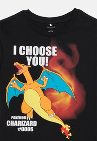 Musta puuvillainen T-paita, jossa on kuvitus Charizardista oranssilla ja sinisellä korostuksella, ja tekstinä "VALITSEN SINUT!" liekkitaustan kanssa.