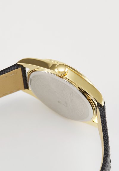 Kuldsetes toonides kell, kus on sile bezels ja tekstureeritud must nahkrihm; ümmargune korpus poleeritud metalltagusega, millel on peeneid detaile.