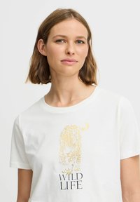 T-shirt en coton blanc avec un graphique doré d'un tigre au-dessus du texte "WILD LIFE" en noir. Manches courtes et encolure ronde.