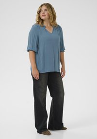 Blue, loose-fitting V-neck blouse with gathered short sleeves, paired with dark flared jeans and light brown shoes. Smooth texture, casual design.