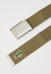Olíva zöld szőtt öv ezüst színű fémből készült csattal, amelyen "LACOSTE" gravírozás és egy kis hímzett krokodil logó található.