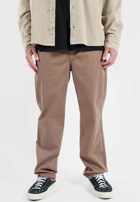Veste en velours côtelé beige à boutons portée par-dessus une chemise noire, associée à un pantalon droit marron et des baskets noires à lacets blancs.