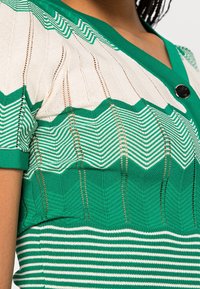Grön och vit randig stickad överdel med ett zick-zackmönster, korta ärmar, en djup V-halsringning och en rund svart knapp. Texturerade tygdetaljer synliga.