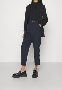 Svart kavaj, marinblå högmidjade byxor med bälte och svarta kraftiga loafers med detaljer i guld. Slätt tyg med skräddarsydd passform.