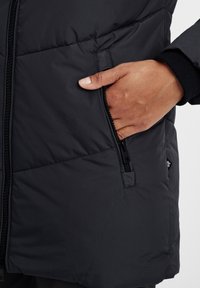 Svart puffjacka med en dragkedjeficka. Materialet verkar slätt med en kviltad textur. Närbild av en hand som håller i fickan.