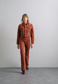 Jachetă scurtă din corduroy maro cu cusături contrastante, asortată cu pantaloni evazați corespunzători. Modelul poartă pantofi negri, fundal simplu.