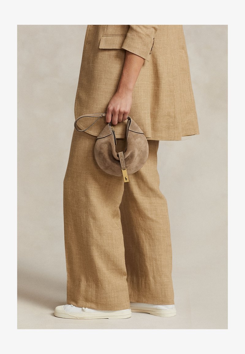 Osoba v béžovom ľanovom obleku a bielych teniskách drží malú okrúhlu taupe semišovú kabelku s remienkom na zápästie a zlatým zipsom.