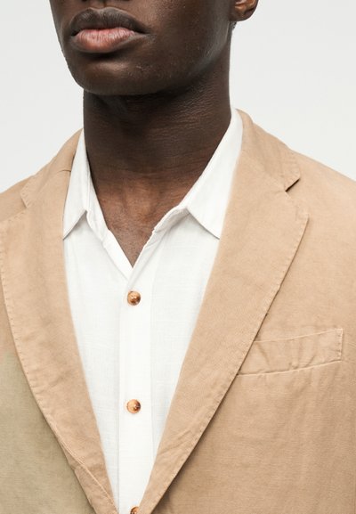 Κοντινό πλάνο προσώπου ατόμου που φοράει μπεζ σακάκι πάνω από λευκό πουκάμισο με κουμπιά από ξύλο, ουδέτερο φόντο.