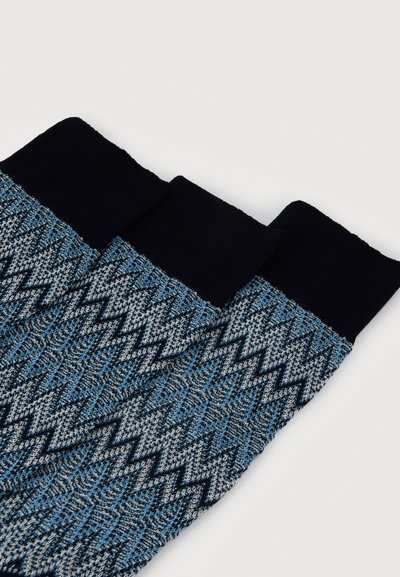 Trois chaussettes bleues et grises à motif en zigzag avec des poignets noirs unis, disposées côte à côte sur un fond blanc.