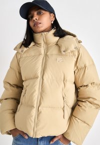 Giacca puffer beige con colletto alto, zip frontale, due tasche laterali con zip e dettagli con bottoni a pressione sulle spalle. Texture liscia e imbottita.
