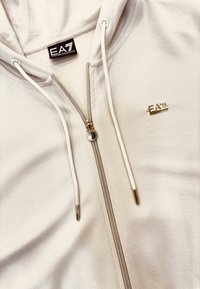 Felpa leggera color beige con zip, realizzata in materiale morbido, dotata di hardware dorato, logo sul petto e cordini sul cappuccio.