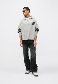 Grijze oversized trui met zwarte accenten, kraagontwerp en logodetail. Gecombineerd met zwarte jeans met brede pijpen en grijze sneakers.