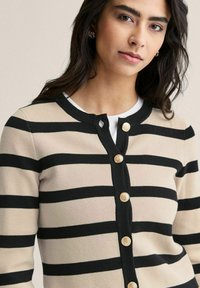 Raidallinen villatakki beige ja musta, jossa on pyöreä kaula-aukko, kultaiset napit ja pehmeä neulepinta. Korostettu vaakasuorilla raidoilla.