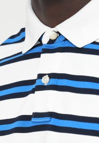 Πόλο μπλούζα σε λευκές, ναυτικές και ανοιχτό γαλάζιες ρίγες. Διαθέτει γιακά με δύο κουμπιά και υφασμένο ύφασμα.