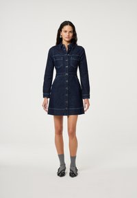 Vrouw staat in een donkerblauwe denim jurk met knopen en lange mouwen, grijze sokken en zwarte schoenen met studs voor een effen witte achtergrond.