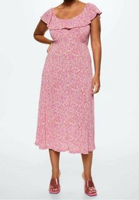 Femme portant une robe midi rose à imprimé floral avec un col à volants et des manches courtes, associée à des sandales à talons transparentes roses.