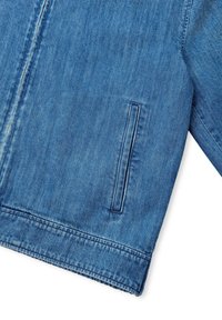 Een deel van een blauwe denimjas met een zijzak en zichtbare stiksels langs de zoom en het ritsgebied op een witte achtergrond.