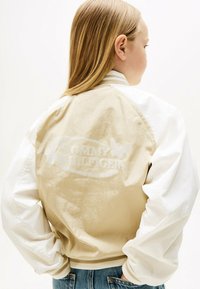 Chaqueta bomber beige y blanca con un acabado brillante, presentando un branding sutil y acentos de mariposa en la parte posterior, puños y cuello acanalados.