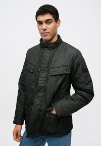 Czarna pikowana kurtka z wysokim kołnierzem, zapięciem na guziki, dwoma przednimi kieszeniami i fakturowanym wzorem. Nosi się ją na niebieskich dżinsach.