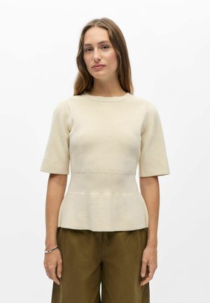 Femme aux longs cheveux bruns portant un haut en maille crème à manches courtes et un pantalon vert olive, debout devant un fond blanc uni.