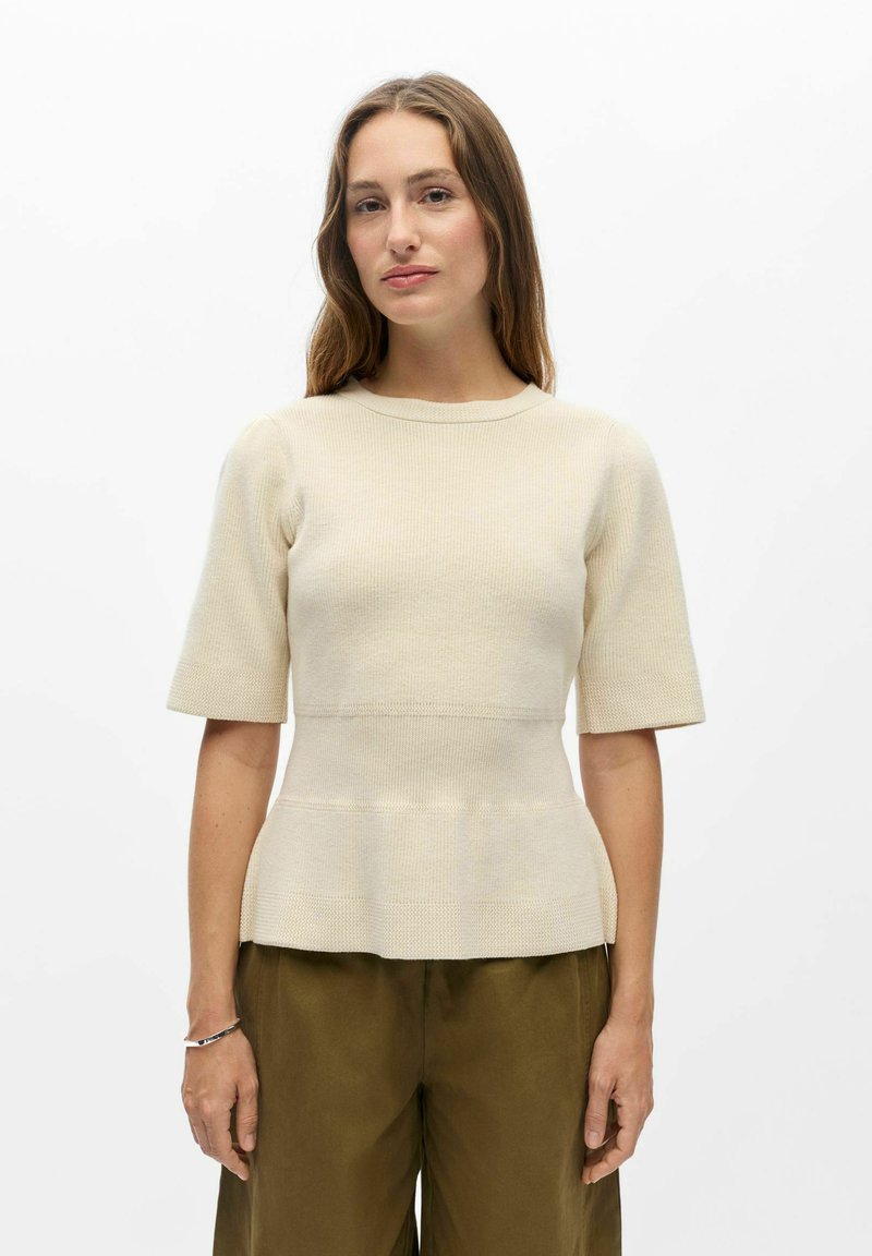 Femme aux longs cheveux bruns portant un haut en maille crème à manches courtes et un pantalon vert olive, debout devant un fond blanc uni.