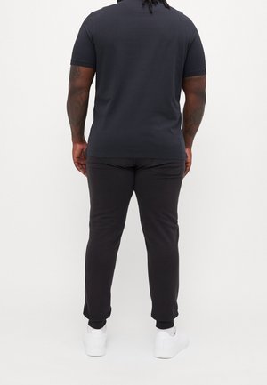 Uomo con tatuaggi che indossa una maglietta nera, pantaloni jogger neri e sneakers bianche, in piedi rivolto di spalle contro uno sfondo bianco semplice.