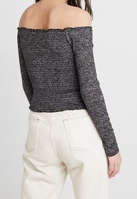 Schulterfreies, langärmliges Oberteil aus dunkelgrauem strukturiertem Strick. Mit einem rüschierten Saum am Ausschnitt, kombiniert mit hellen, hochgeschnittenen Jeans.