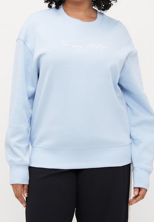 Kobieta ubrana w jasnoniebieską bluzę z długim rękawem z wyhaftowanym na piersi napisem „Tommy Hilfiger” oraz czarne spodnie.