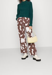 Persoon in een groen geribbeld trui, bruine en witte koeienvlek brede broek, witte chunky laarzen, met een lichtgele handtas.