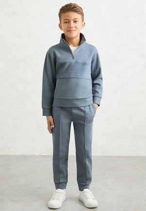 Gaiši zils fliša džemperis ar rāvējslēdzēju un teksturētu joslu, kopā ar atbilstošām šaurām biksēm. Abām drēbēm ir elastīgi aproces un sānu kabatas.