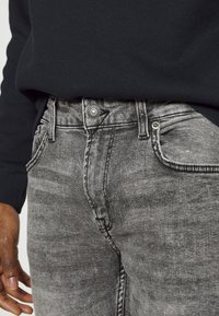 Jeans de mezclilla gris con una textura deslavada, cinco bolsillos y herrajes de metal. Un suéter negro es parcialmente visible sobre la cinturilla.